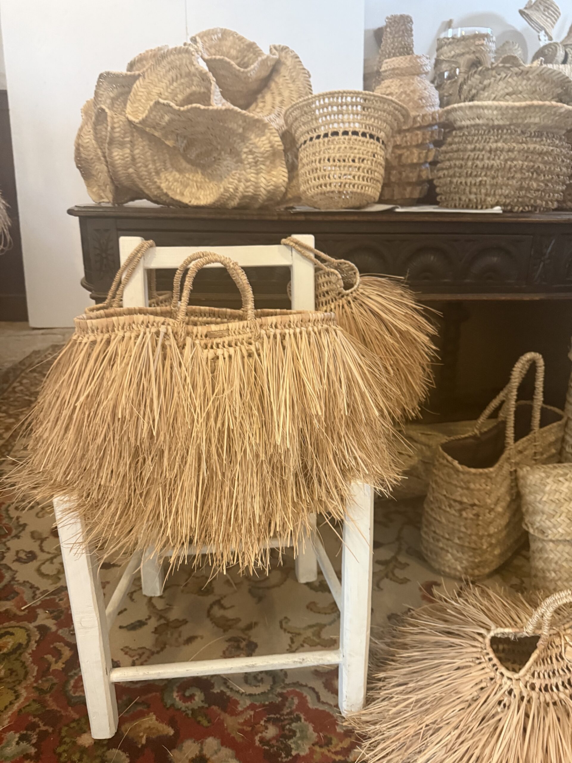 A fringed palm tote resting beside a collection of finished baskets in the atelier
