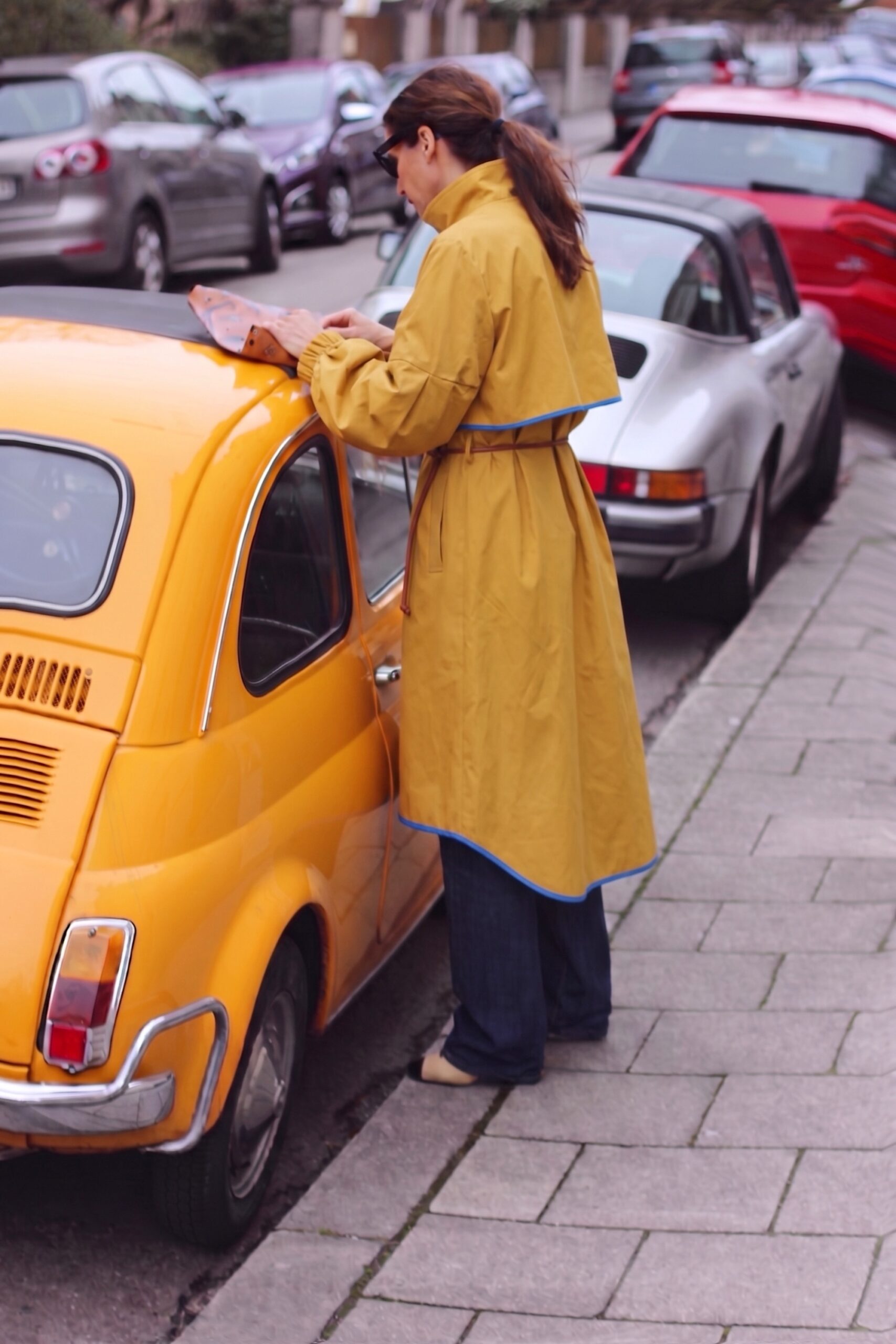 Woman in a mustard trench coat with blue trim standing beside a vintage yellow Fiat