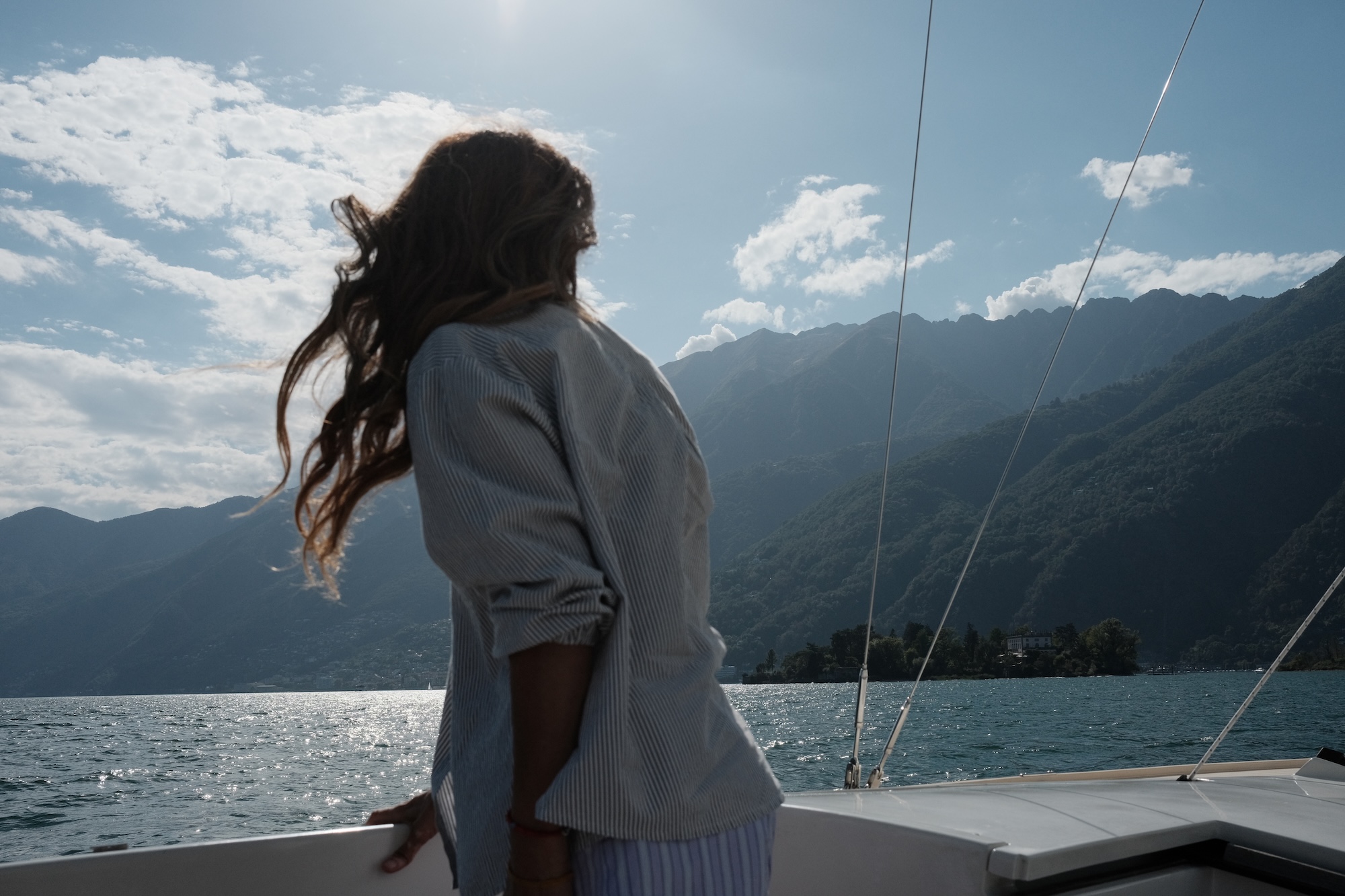 A guest on the bow of the hotel's sailboat, hair in the wind, mountains along the shore